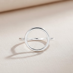 Puede incluir: Un anillo minimalista de plata con un diseño de círculo hueco. El anillo se presenta sobre un fondo neutro, resaltando su estética simple y moderna. La banda del anillo es delgada y el círculo está centrado, creando un aspecto limpio y elegante. Joyería.