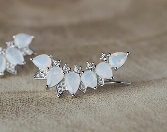 May include: A pair of silver ear cuffs with white opal stones. The ear cuffs are shaped like a crescent moon and have a delicate design.