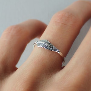 May include: A silver ring featuring a feather design. The ring is worn on a finger.