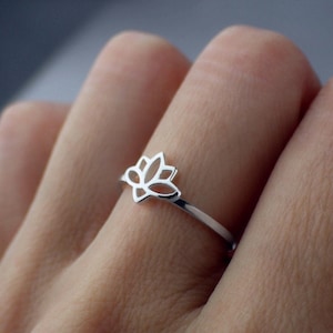 Può includere: Un anello d'argento con un delicato design a forma di fiore di loto. L'anello è indossato su un dito.