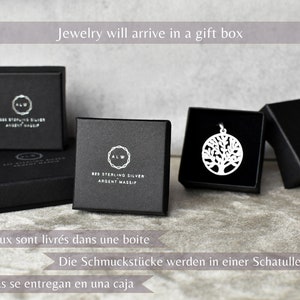 May include: Four black boxes with silver lettering that reads "925 Sterling Silver Argent Massif" and "ALW". The boxes are on a gray surface. One box contains a silver tree of life pendant.