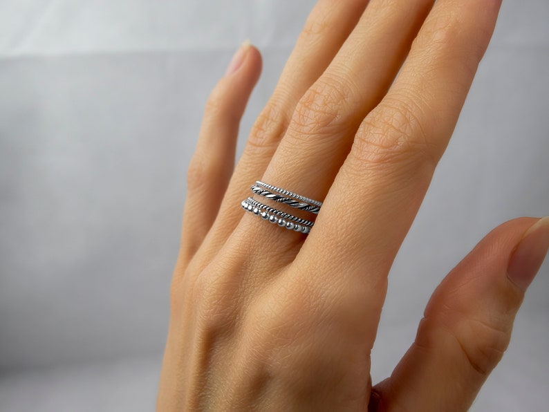 May include: A set of three silver rings stacked on a finger. The top ring is a thin band, the middle ring is a twisted band, and the bottom ring is a band with small beads.
