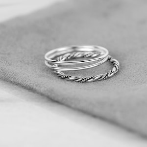May include: Three silver rings, one with a twisted design, stacked on a gray fabric surface.