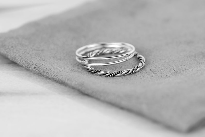 May include: Three silver rings, one with a twisted design, stacked on a gray fabric surface.