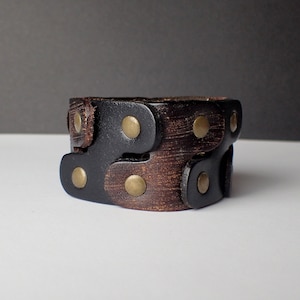 Peut inclure: Un bracelet en cuir marron et noir avec des clous en laiton. Le bracelet est composé de deux couches de cuir, une marron et une noire, et est sécurisé par une boucle.