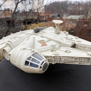 Może przedstawiać: Szczegółowy, kremowy model statku kosmicznego Millennium Falcon. Model posiada skomplikowane panele, kokpit z niebieskimi oknami i charakterystyczną antenę radarową. Zabawka kolekcjonerska z serii Gwiezdne Wojny.