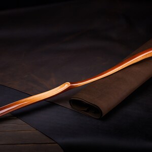May include: A wooden shoehorn with a curved handle and a smooth, polished finish. The shoehorn is resting on a brown leather surface.