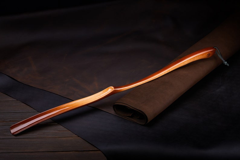 May include: A wooden shoehorn with a curved handle and a smooth, polished finish. The shoehorn is resting on a brown leather surface.