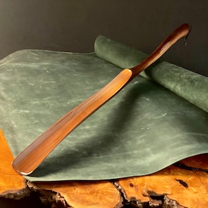 May include: A wooden shoehorn with a curved handle, resting on a green leather surface. The shoehorn is made of dark wood and has a smooth, polished finish.