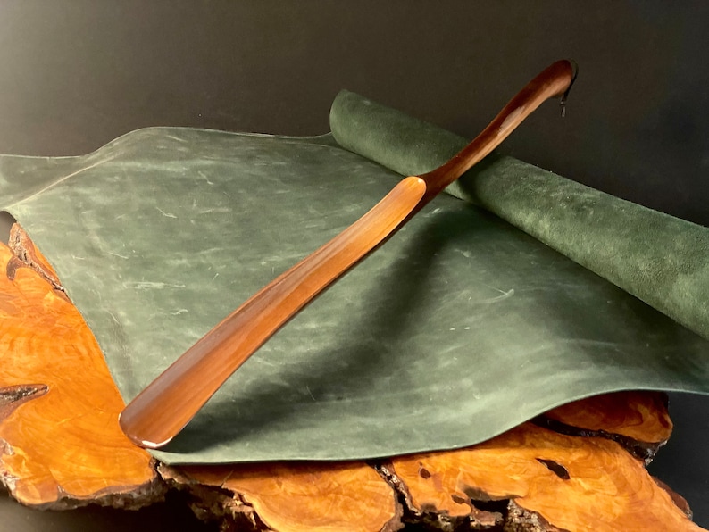 May include: A wooden shoehorn with a curved handle, resting on a green leather surface. The shoehorn is made of dark wood and has a smooth, polished finish.