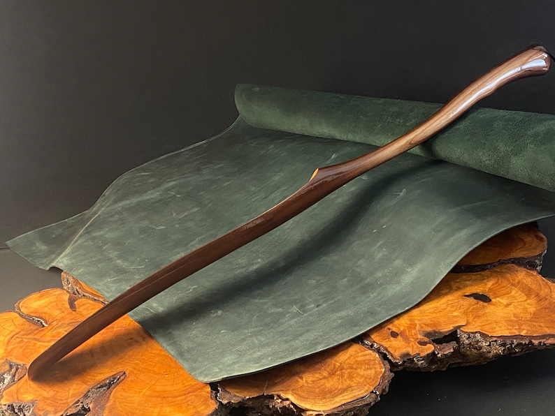 May include: A long, curved wooden shoe horn with a dark brown finish. The horn is resting on a green suede surface, which is on top of a wooden base.