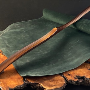 May include: A long, wooden shoehorn with a curved handle. The shoehorn is made of dark wood with a lighter wood grain running through it. The shoehorn is resting on a piece of green suede and a wooden surface.