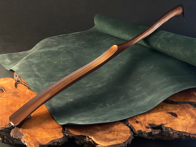 May include: A long, wooden shoehorn with a curved handle. The shoehorn is made of dark wood with a lighter wood grain running through it. The shoehorn is resting on a piece of green suede and a wooden surface.