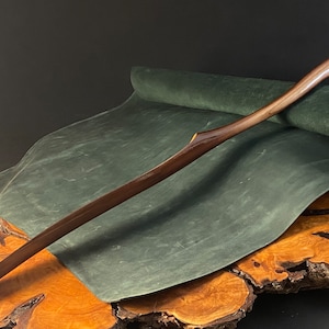 May include: A long, curved wooden shoe horn with a dark brown finish. The horn is resting on a green suede surface, which is on top of a wooden base.