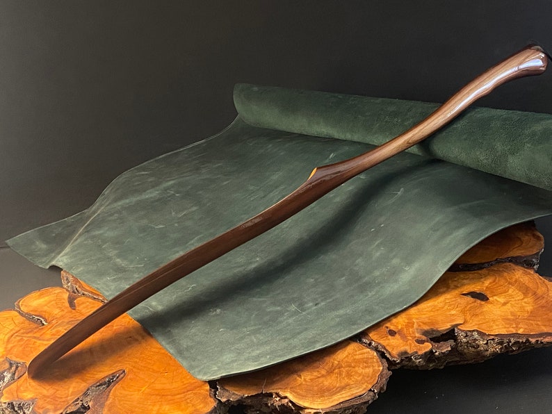 May include: A long, curved wooden shoe horn with a dark brown finish. The horn is resting on a green suede surface, which is on top of a wooden base.