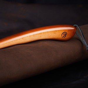 May include: A wooden shoe horn with a brown leather strap. The shoe horn has a smooth, polished finish and a small engraved logo on the handle.