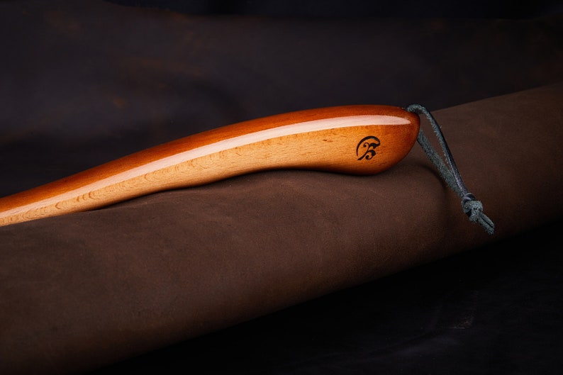 May include: A wooden shoe horn with a brown leather strap. The shoe horn has a smooth, polished finish and a small engraved logo on the handle.