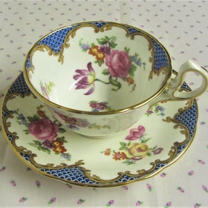 May include: A white teacup and saucer with a blue and gold floral pattern. The cup and saucer have a scalloped edge and a gold trim.