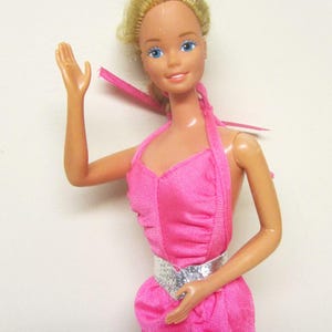 May include: A vintage doll with blonde hair and blue eyes, dressed in a bright pink halter dress with a silver belt. The doll is posed with one arm raised, and the dress has a ruffled design.