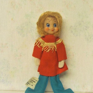 Peut inclure: Une poupée vintage aux cheveux blonds, aux yeux bleus et aux joues roses. Elle porte une robe rouge avec une bordure dorée et un pantalon bleu. La poupée mesure environ 25 cm de haut.