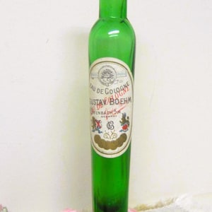May include: A tall, slender, emerald green glass bottle with a long neck. The bottle features a white paper label with the text "EAU DE COLOGNE GUSTAV BOEHM OFFENBACH A.M. GERMANY" and a smaller label on the neck reading "AV BOEHM OFFENBACH A.M."