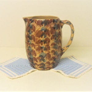 May include: A brown ceramic jug with a blue and brown floral pattern. The jug has a handle and a rounded body.