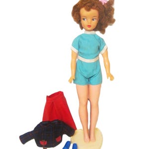 May include: A vintage doll with brown curly hair, wearing a blue and white outfit, standing on a cream-colored base. The doll is holding a pink flower in her hair. The doll is also wearing a red skirt, a plaid jacket, and blue shoes.