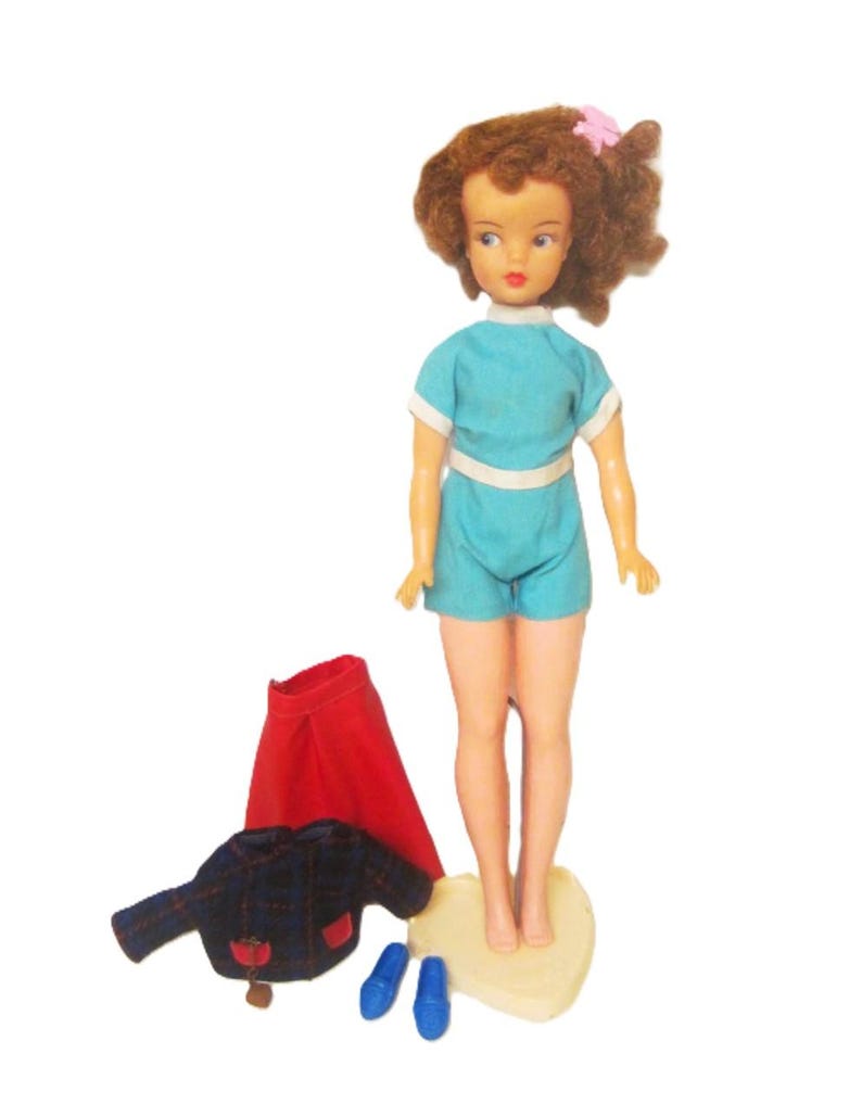 May include: A vintage doll with brown curly hair, wearing a blue and white outfit, standing on a cream-colored base. The doll is holding a pink flower in her hair. The doll is also wearing a red skirt, a plaid jacket, and blue shoes.