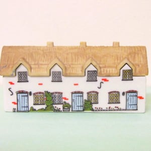 Peut inclure: Une figurine de cottage en céramique avec un toit de chaume beige texturé et une façade blanche. L'extérieur présente trois portes bleues, plusieurs fenêtres à carreaux et des plantes grimpantes vertes peintes. Des briques rouges sont visibles.