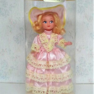 Puede incluir: Una mu&ntilde;eca vintage que lleva un vestido rosa con ribetes de encaje amarillo y un sombrero amarillo. La mu&ntilde;eca est&aacute; en un recipiente de pl&aacute;stico transparente con una etiqueta que dice "COLLECTION" y "U.S.A. ETATS-UNIS".