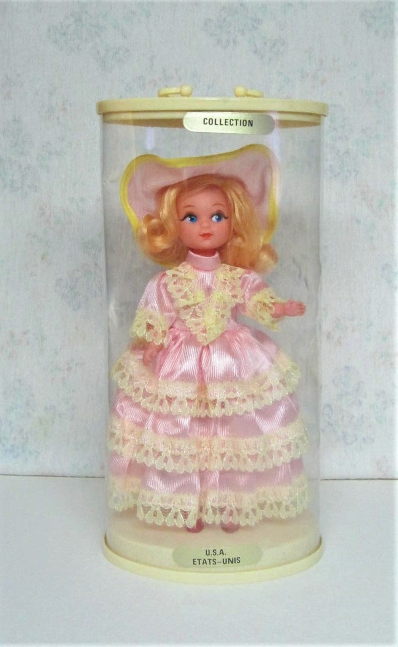 Puede incluir: Una mu&ntilde;eca vintage que lleva un vestido rosa con ribetes de encaje amarillo y un sombrero amarillo. La mu&ntilde;eca est&aacute; en un recipiente de pl&aacute;stico transparente con una etiqueta que dice "COLLECTION" y "U.S.A. ETATS-UNIS".