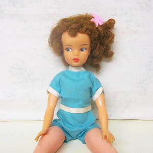 May include: A vintage doll with brown curly hair, wearing a blue and white one-piece outfit. The doll has blue eyes and is wearing a pink flower in her hair.