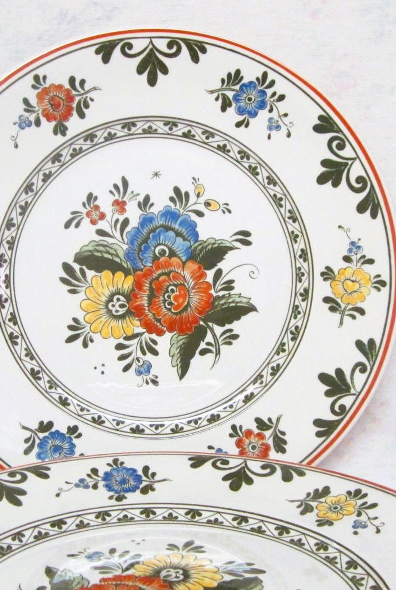 May include: Two white ceramic dinner plates with a floral design. The plates have a green and red border with a white center. The center of the plate features a bouquet of yellow, red, and blue flowers with green leaves.