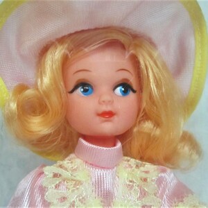 Puede incluir: Una mu&ntilde;eca vintage con cabello rubio, vestida con un vestido rosa con encaje amarillo y un sombrero rosa y amarillo.