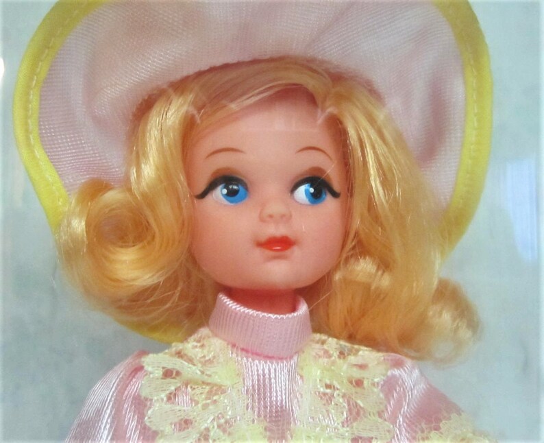 Puede incluir: Una mu&ntilde;eca vintage con cabello rubio, vestida con un vestido rosa con encaje amarillo y un sombrero rosa y amarillo.
