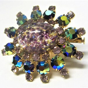 Peut inclure: Une broche dor&eacute;e avec une grande pierre centrale violette iris&eacute;e entour&eacute;e de petites pierres bleues, vertes et violettes iris&eacute;es.