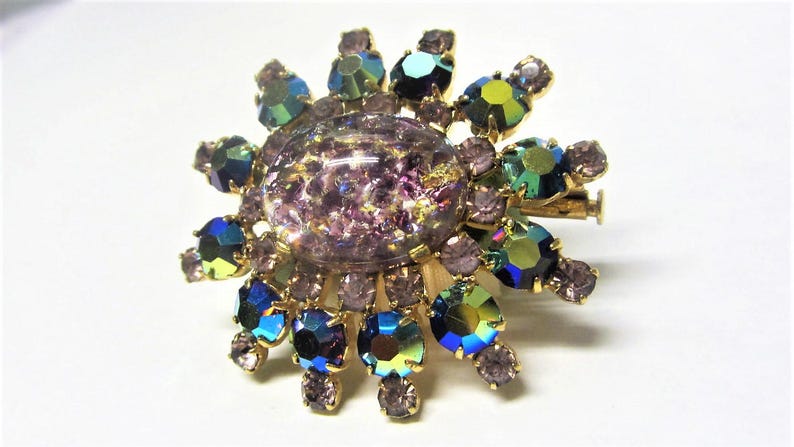 Peut inclure: Une broche dor&eacute;e avec une grande pierre centrale violette iris&eacute;e entour&eacute;e de petites pierres bleues, vertes et violettes iris&eacute;es.
