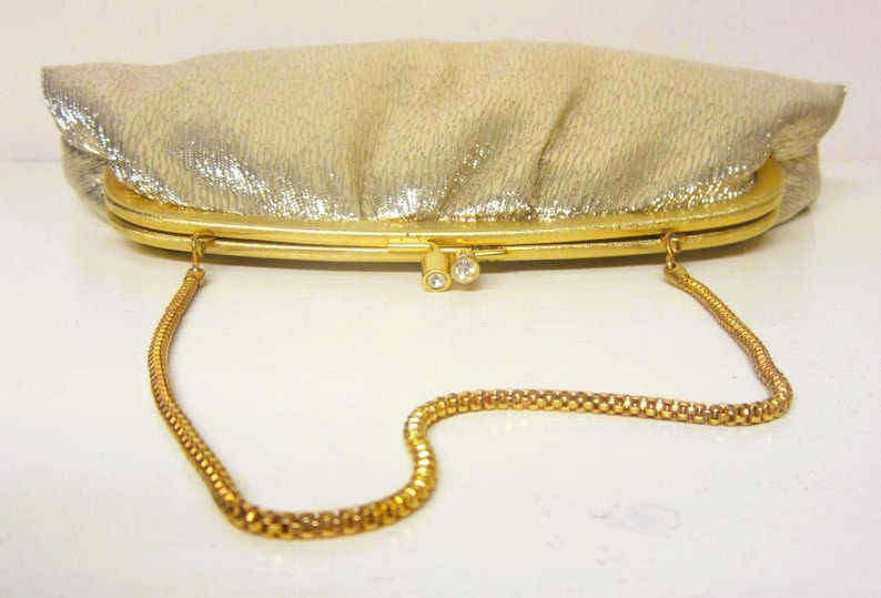 K&ouml;nnte beinhalten: Eine goldene Vintage-Clutch mit strukturierter, schimmernder Oberfl&auml;che. Die Clutch hat einen goldenen Rahmen, einen Verschluss mit zwei kleinen Strasssteinen und einen zarten goldenen Kettenriemen. Ein klassisches Accessoire f&uuml;r besondere Anl&auml;sse.