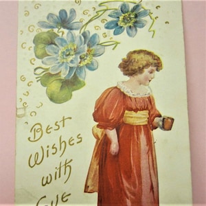 Puede incluir: Una postal vintage con una ni&ntilde;a peque&ntilde;a con un vestido rojo sosteniendo una taza. La ni&ntilde;a est&aacute; rodeada de flores azules y hojas verdes. El texto "Mejores deseos con amor" est&aacute; escrito en letras doradas.