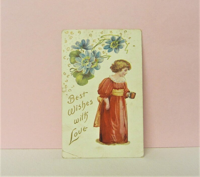 May include: Vintage postcard featuring a girl in a red dress holding a cup, with blue flowers and green leaves in the background. The text "Best Wishes with Love" is written in gold script.