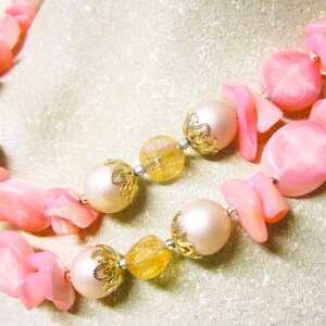 May include: A double strand necklace with pink and white beads. The beads are made of plastic and have a glossy finish. The necklace is accented with gold-colored metal beads.
