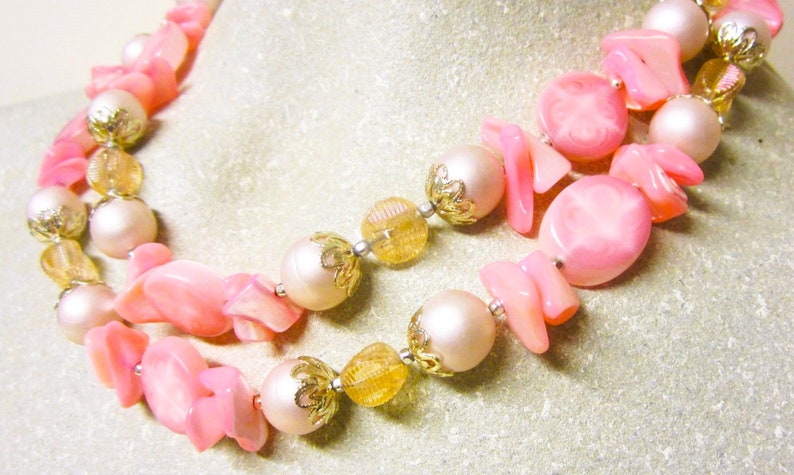 May include: A double strand necklace with pink and white beads. The beads are made of plastic and have a glossy finish. The necklace is accented with gold-colored metal beads.