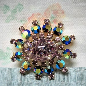 Peut inclure: Une grande broche en strass rose iris&eacute; avec un groupe de plus petits strass bleus, verts et violets autour du centre.