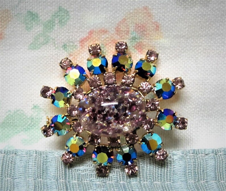 Peut inclure: Une grande broche en strass rose iris&eacute; avec un groupe de plus petits strass bleus, verts et violets autour du centre.