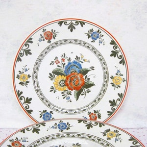 May include: Two white ceramic plates with a floral design. The plates have a green, red, yellow, and blue floral pattern with a white background. The plates have a red rim.