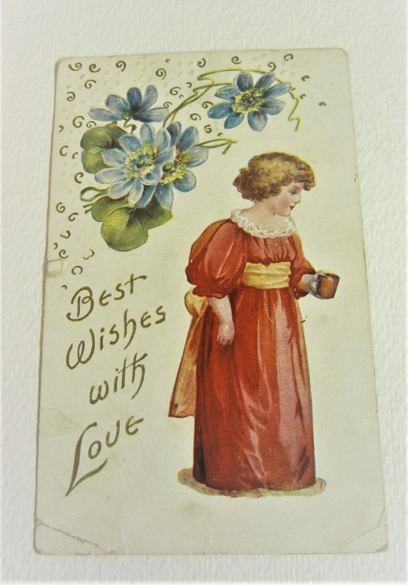 Puede incluir: Una postal vintage con una ni&ntilde;a joven con un vestido rojo sosteniendo una taza. La ni&ntilde;a est&aacute; de pie frente a un dise&ntilde;o floral con flores azules y hojas verdes. El texto "Mejores deseos con amor" est&aacute; escrito en cursiva en la postal.