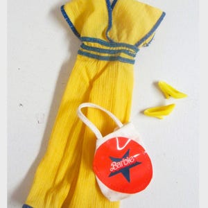 Peut inclure: Une robe de poupée jaune avec une bordure bleue, un sac à main blanc et rouge avec le mot "Barbie" et deux chaussures de poupée jaunes. La robe a une encolure à volants et une taille ajustée. Le sac a un motif d'étoile.