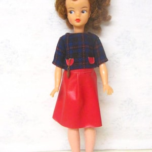 May include: A vintage doll with brown curly hair, wearing a blue and black plaid top with red accents, a red skirt, and blue shoes.