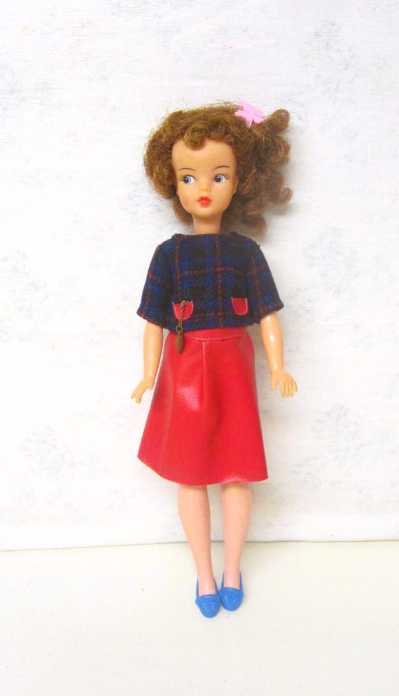 May include: A vintage doll with brown curly hair, wearing a blue and black plaid top with red accents, a red skirt, and blue shoes.