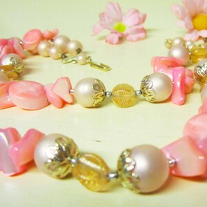 May include: A pink and white beaded necklace with gold accents. The necklace features a variety of pink and white beads, including some with a pearlescent finish. The necklace is strung on a gold chain.